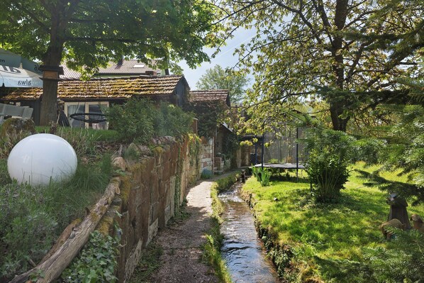 A narrow path leading to a rustic building with a thatched roof, surrounded by greenery and a small stream.