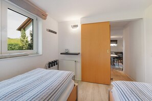 A bedroom with two beds, a window, a radiator, and a wooden door leading to a kitchen.