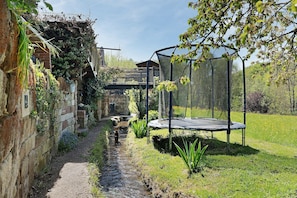 Ein schmaler Pfad führt zu einem Trampolin in einem Garten mit einer Backsteinmauer und viel Grün.