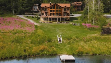 Une grande cabane en bois dotée d’une terrasse, située au bord d’un lac avec un quai et un bateau.