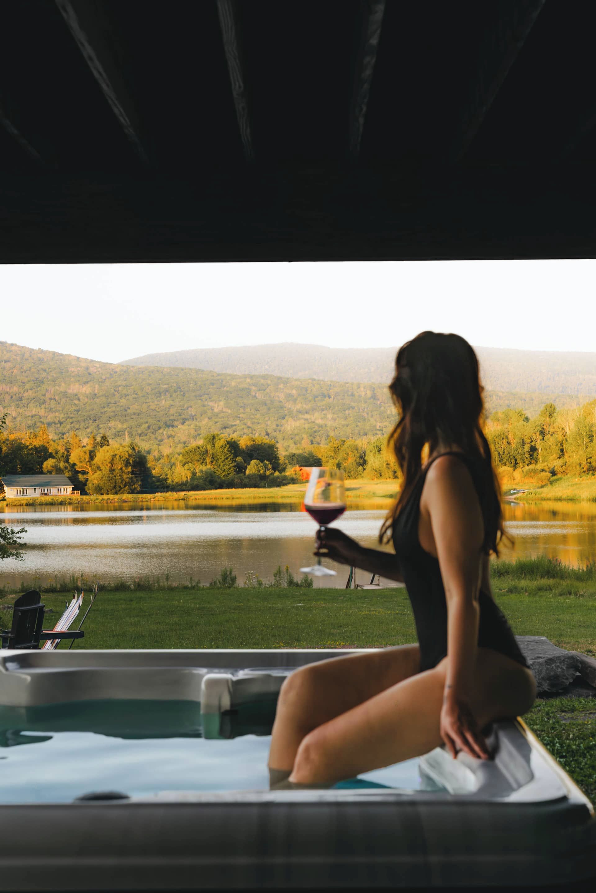A person sitting on a bench by a lake with a view of a house and hills in the background.
