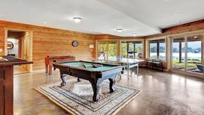 A spacious indoor area with a pool table, foosball table, and ping pong table. There are large windows offering views of a lake. - The Hunter Chalet (Hunter)