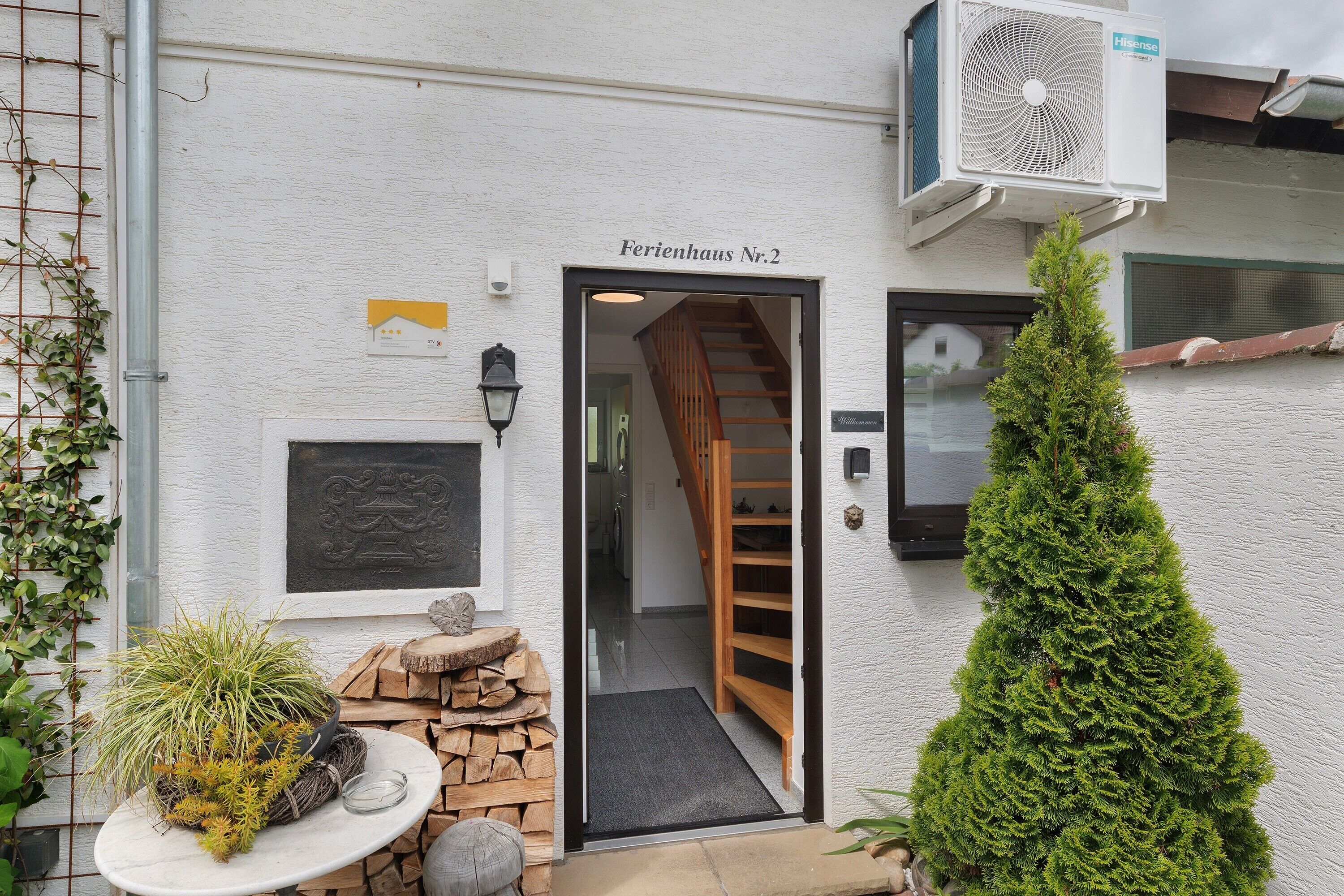 Une entrée d’immeuble avec un escalier, une petite plante en pot et un grand conifère.
