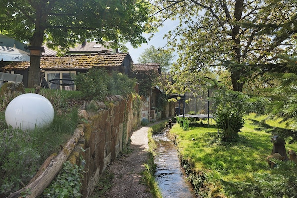 A narrow path leading to a rustic building with a thatched roof, surrounded by greenery and a small stream.