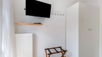 A room with a mounted TV, a radiator, a wooden shelf, a small wooden stool, and a white wardrobe.