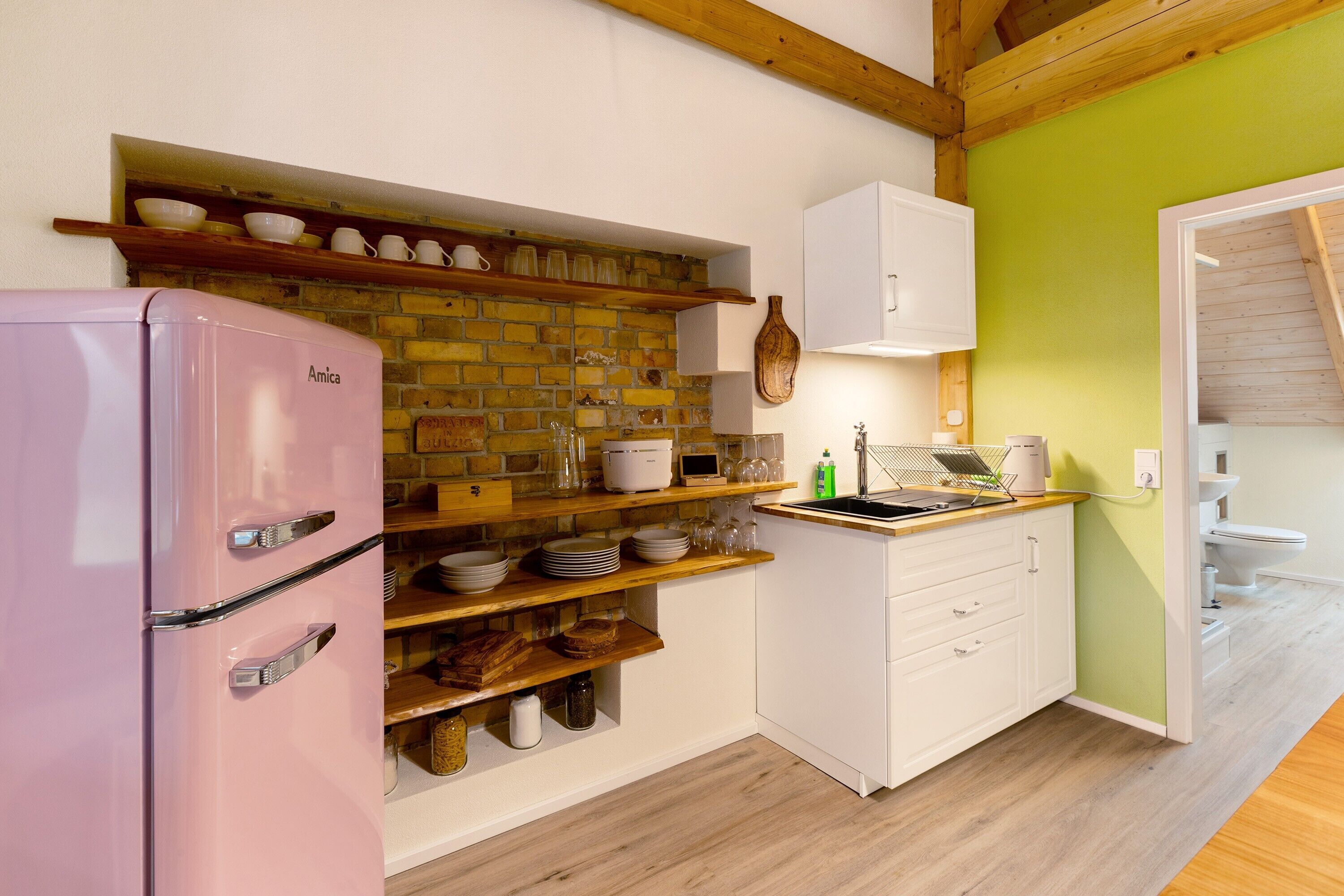 A kitchen with a pink refrigerator, wooden shelves, white cabinets, and a brick wall.