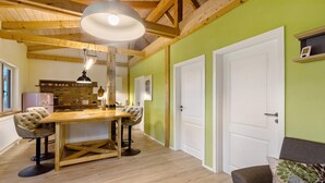 A dining area with a wooden table and chairs, a kitchen in the background, and a green wall with a white door. - Apartment 'Fewo Am Flämingrand' with Shared Garden, Balcony and Wi-Fi (Zahna-Elster)
