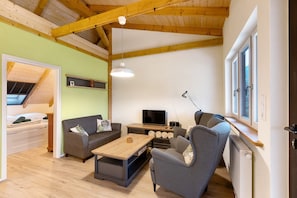 A cozy living room with a sofa, armchair, coffee table, TV, and wooden beams on the ceiling.