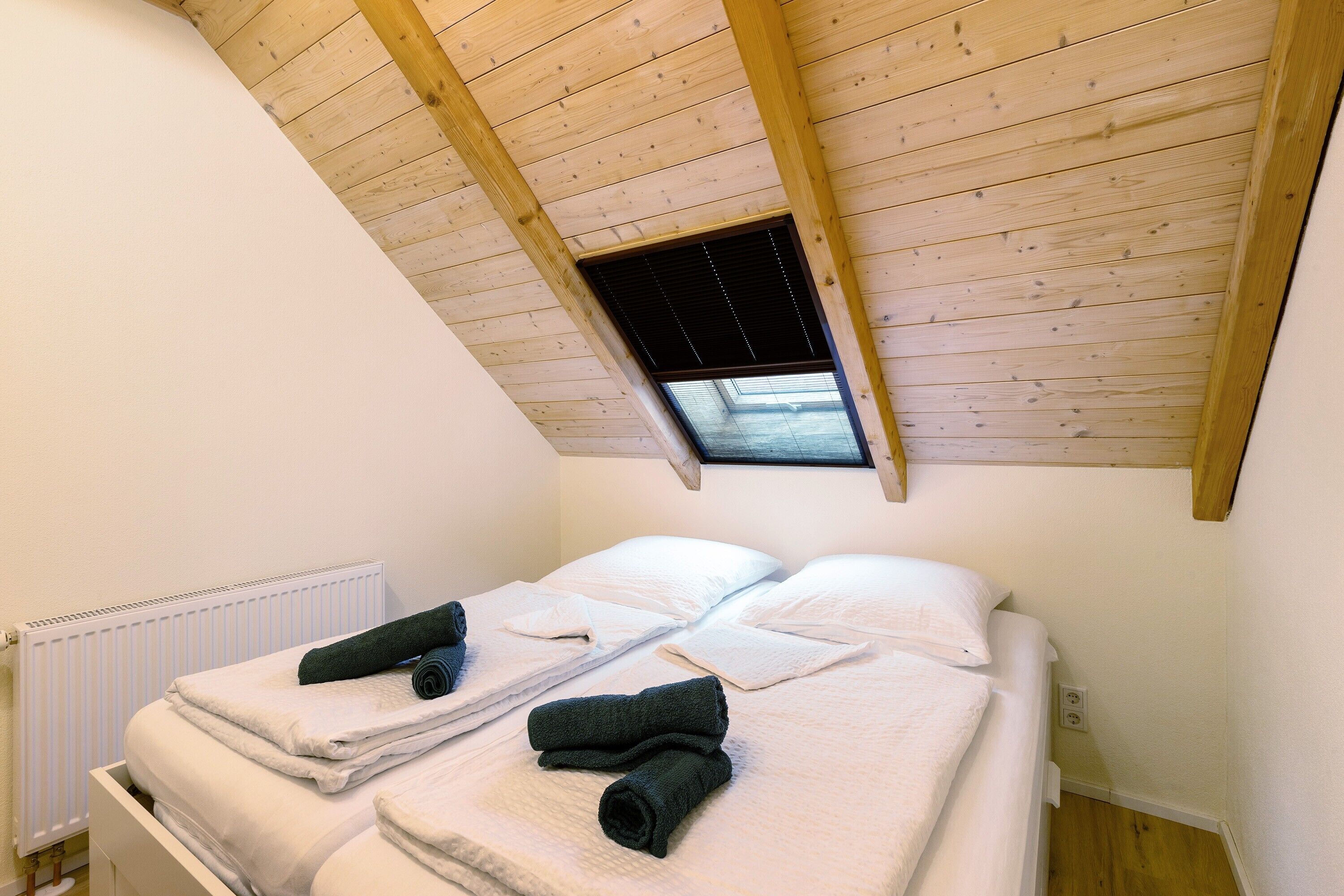 Een slaapkamer met twee bedden, houten balken in het plafond, een dakraam en een witte radiator.
