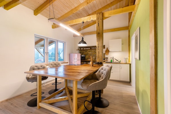 A dining area with a wooden table and chairs, a hanging light fixture, and a view of the kitchen in the background.