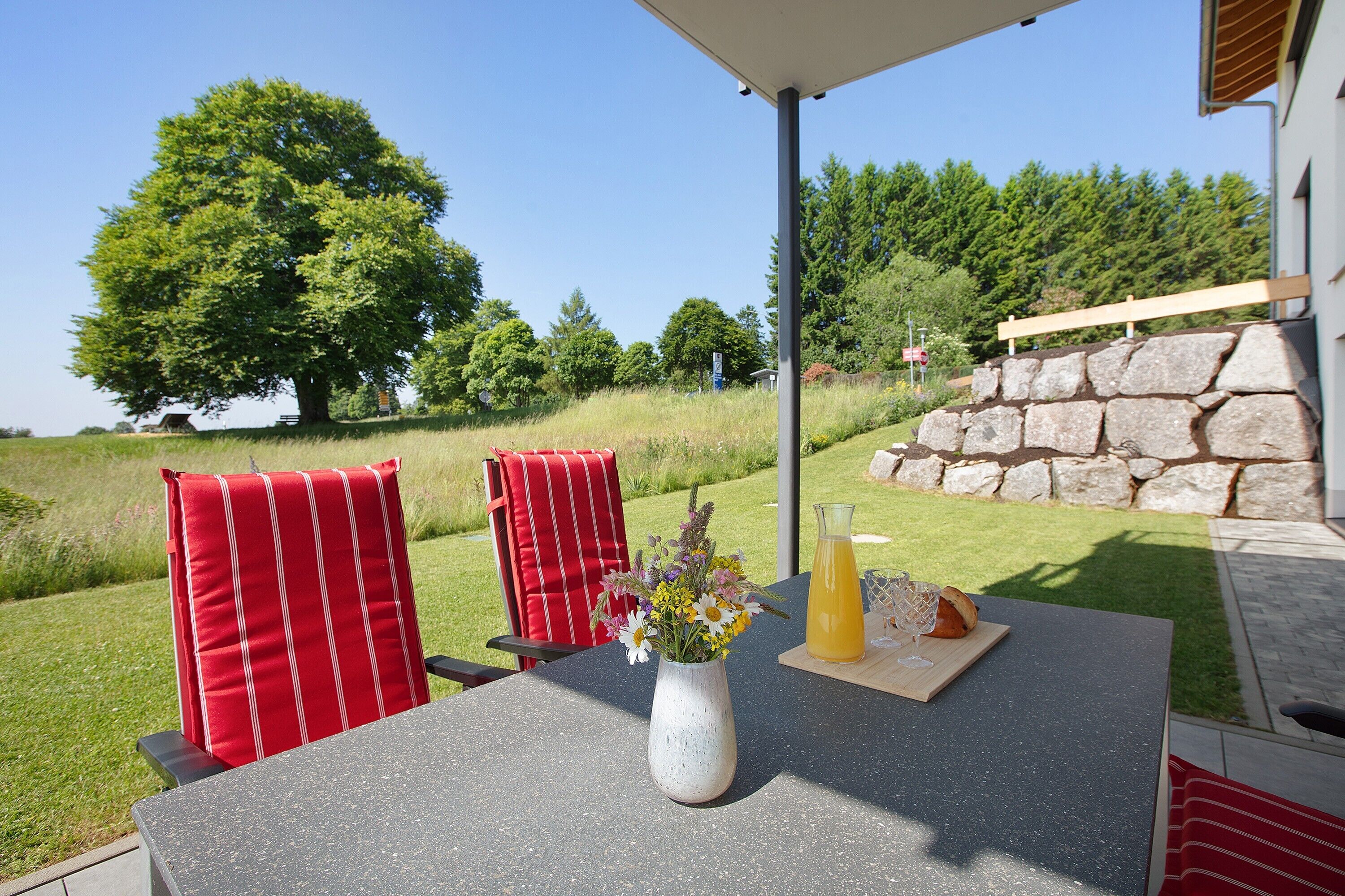 Een buitentafel met een bloevas, een glas sinaasappelsap en een brood op een patio met rood gestreepte stoelen.