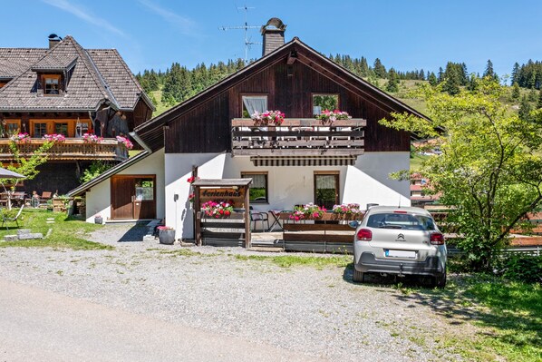 Une maison en bois à deux étages, dotée d’un balcon, avec une voiture garée devant et une pancarte indiquant « Ferienhaus ».