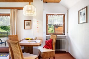 Una habitación con una mesa de comedor de madera, sillas de mimbre, un radiador, una ventana con vistas a follaje y un cuadro colgado en la pared.