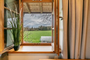 Una finestra con vista su un campo verde e alberi, una pianta in vaso sul davanzale e un soffitto in legno.