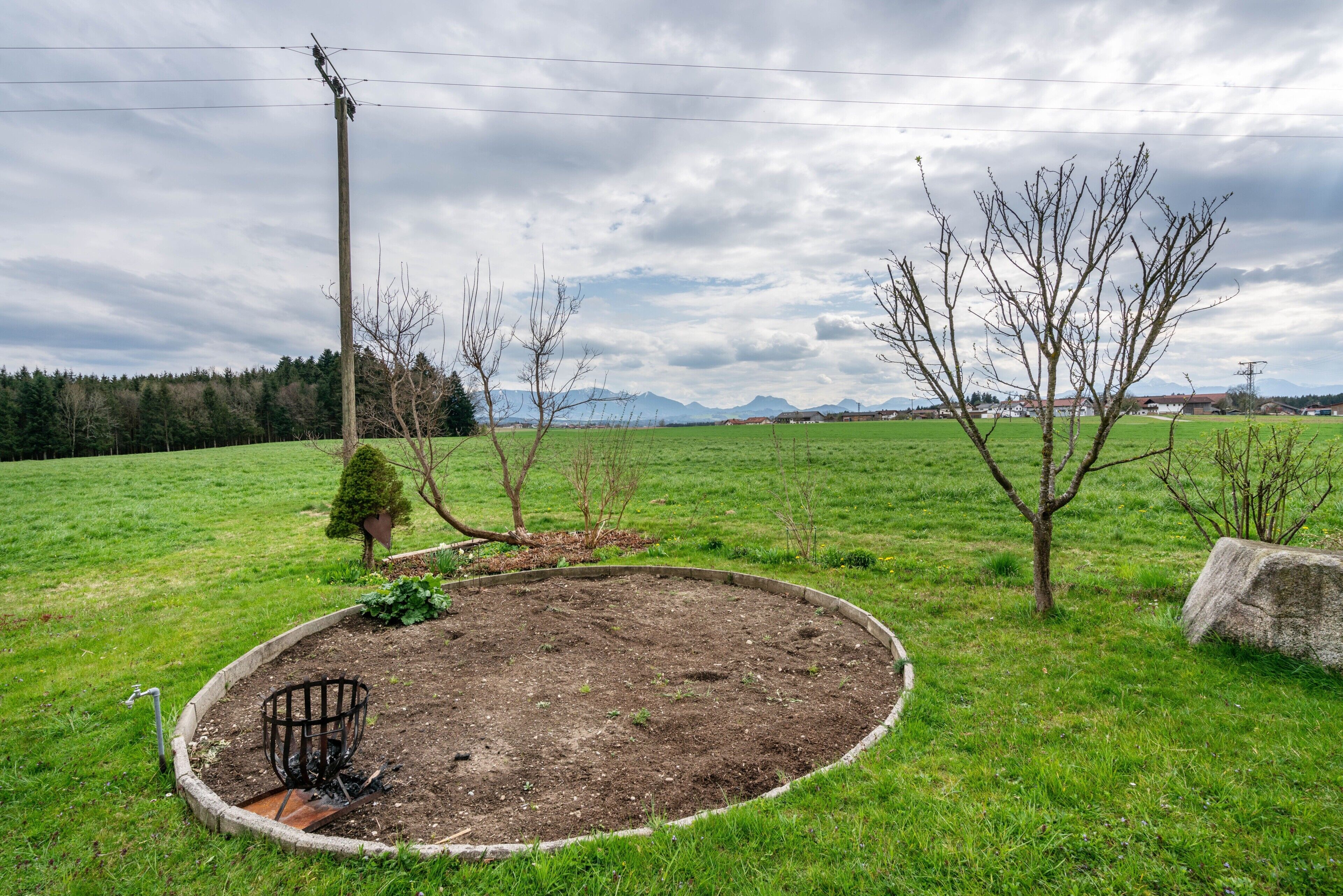 Een ronde bloembed met een metalen plantenbak, een steen en een boom in een grasveld, met een hoogspanningskabel en op de achtergrond bergen.