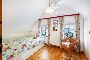 Ein Schlafzimmer mit Bett, Sessel, einem Fenster mit Vorhängen, Heizkörper und einer Wandleuchte.