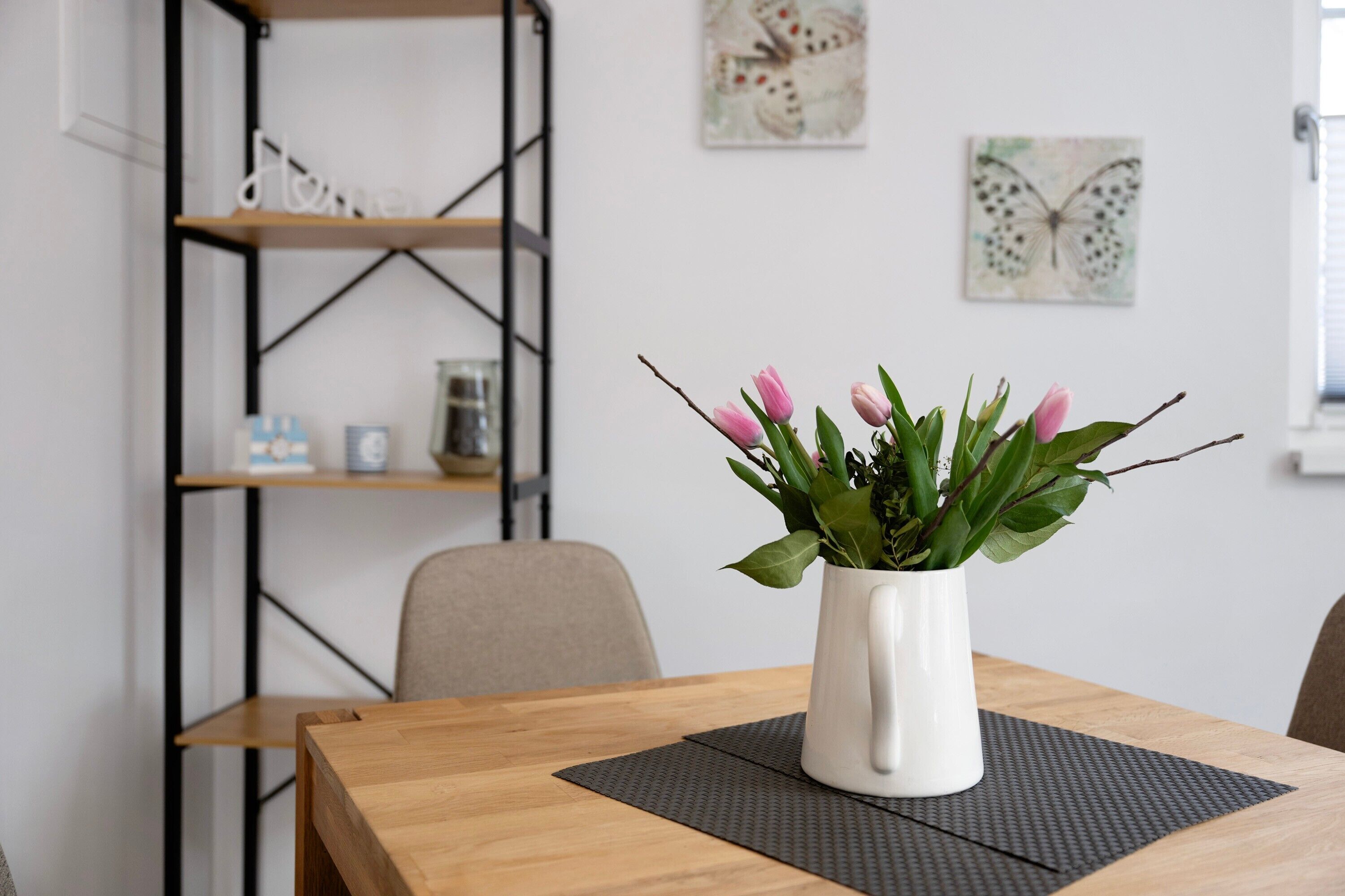 Una mesa de comedor con un jarrón blanco lleno de flores rosas, una silla de madera y una base para mantel negra.