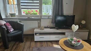 A living room with a television, a sofa, a small table with a heart-shaped decoration, and a window with blinds.