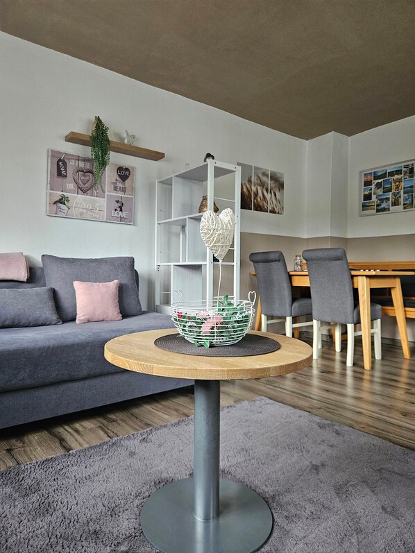 Una sala de estar moderna con un sofá gris, una mesa redonda de madera con una cesta blanca, sillas grises y un estante blanco.