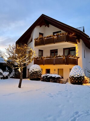 Um edifício de dois andares com varanda, chão coberto de neve e luzes decorativas.