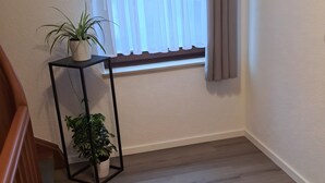 Una stanza con pavimento in legno, una finestra con tende leggere e due piante in vaso su un supporto nero.