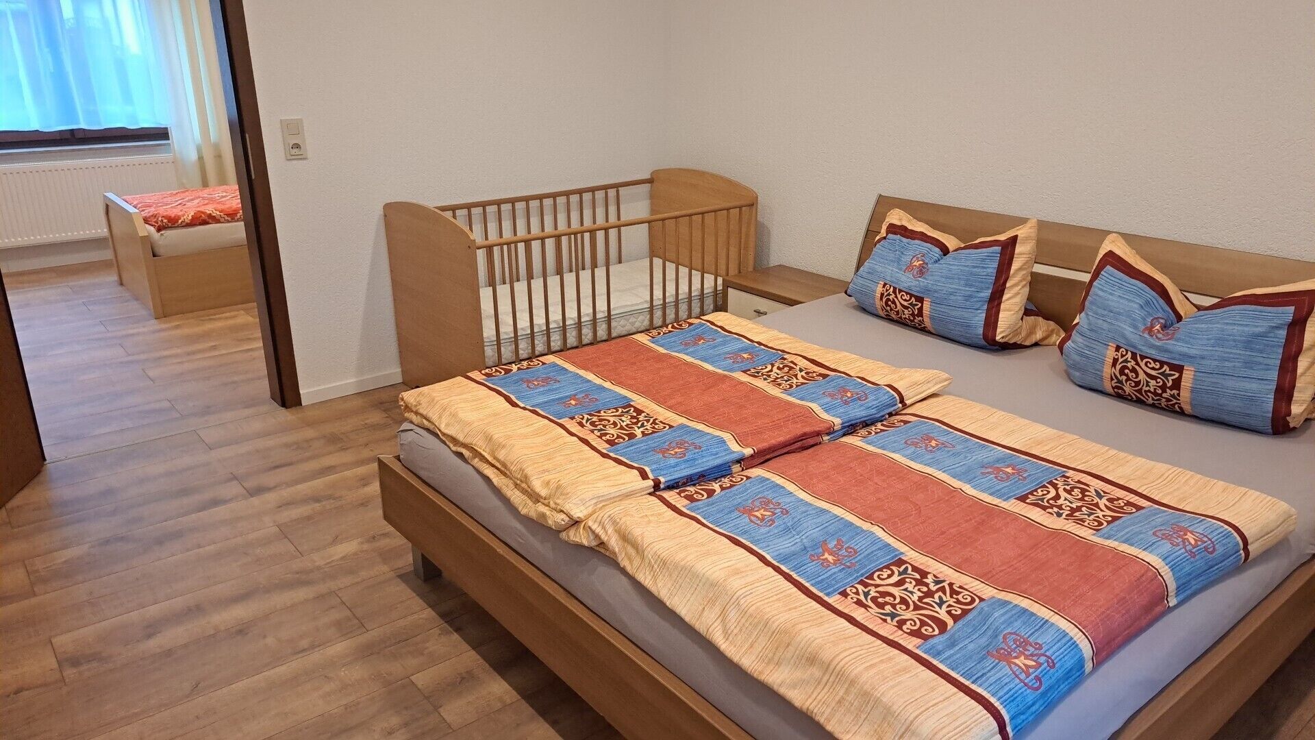 Una habitación con una cama, una cuna de madera y un sofá con cojines estampados.