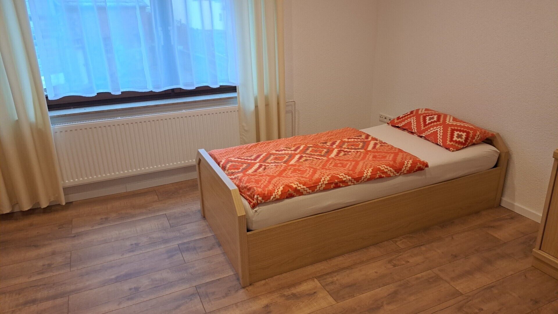 Ein kleines Zimmer mit einem Einzelbett, einem Holrahmen, einem Fenster mit durchscheinenden Vorhängen und einem Heizkörper darunter.