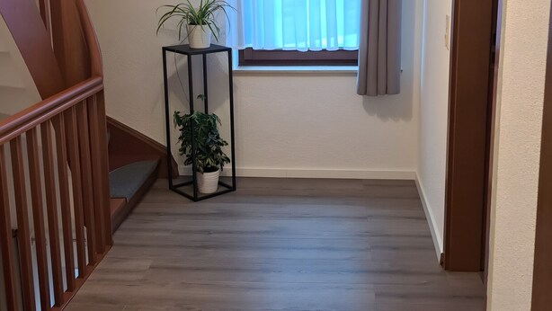Un corridoio con pavimento in legno, una scala, una finestra con tende e una pianta in vaso.