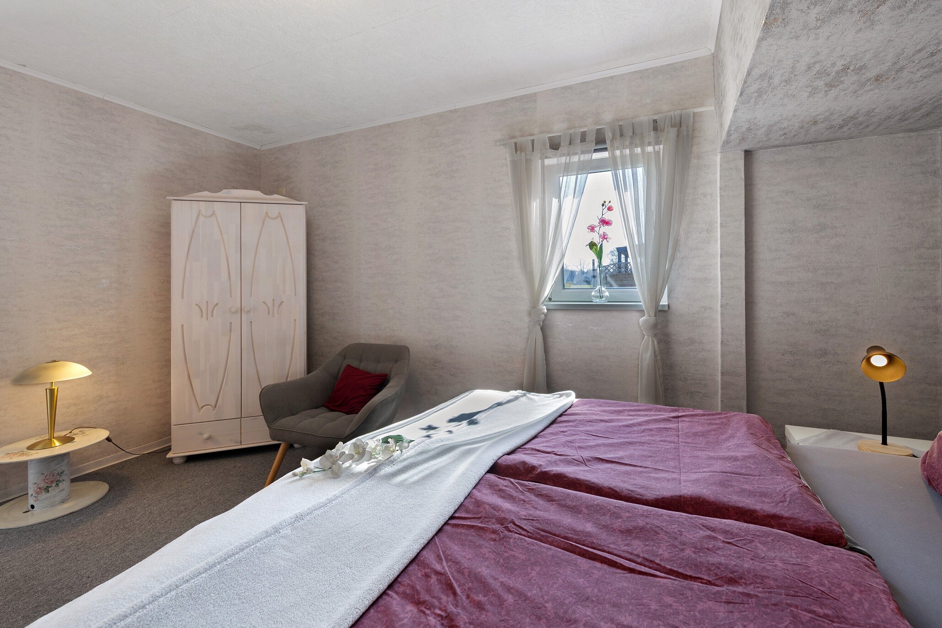Ein Schlafzimmer mit Bett, Sessel, Kleiderschrank, einem Fenster mit Vorhängen und einer Nachttischlampe.