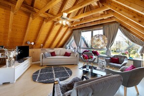 A spacious loft-style living area with a high ceiling, wooden beams, a sofa set, a coffee table, and a television.