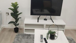 A living room with a flat-screen TV on a white stand, a small potted plant, a remote control, and a fluffy rug. - 1 Bed Studio - Central, Free Parking (London)