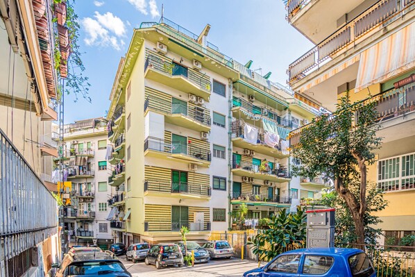A multi-story residential building with balconies, a parking area, and a street view.