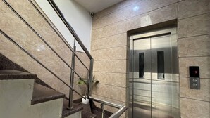 Un ascensor moderno con detalles metálicos, junto a una escalera con barandillas de metal y paredes con mosaicos de mármol.