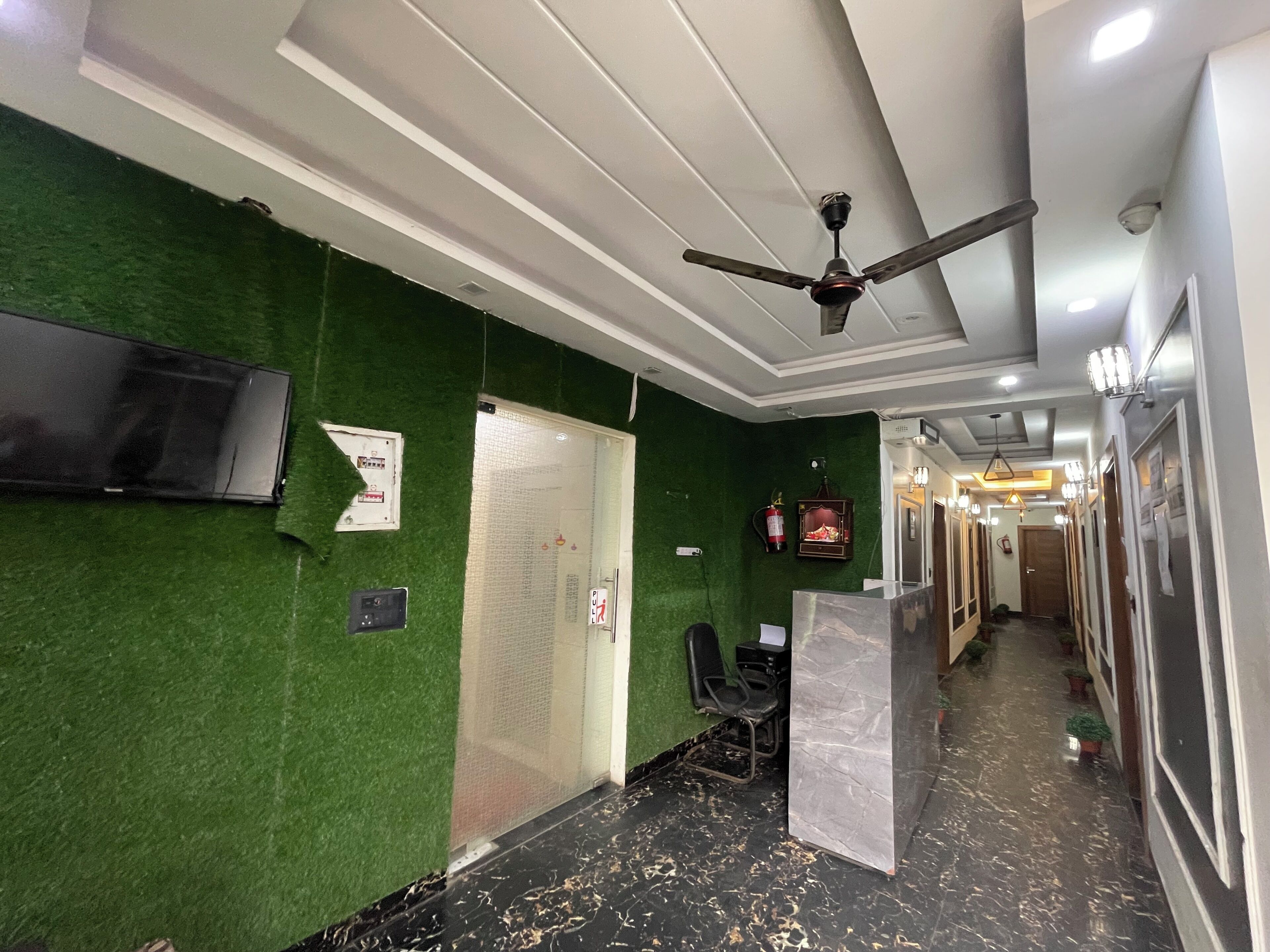 Um hall de hotel com parede verde, um ventilador de teto, uma televisão fixada na parede e uma mesa de recepção.
