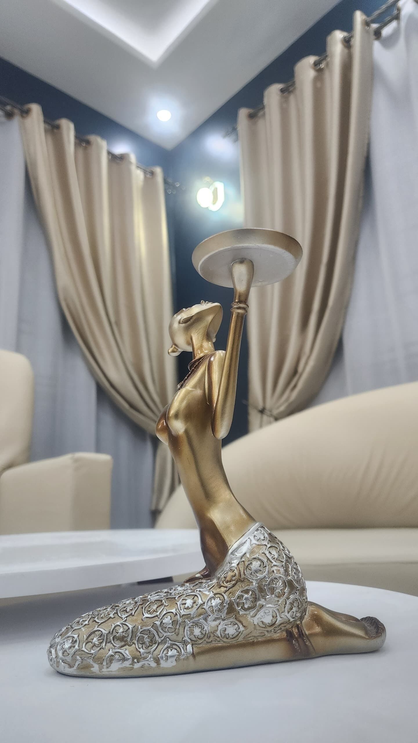 Una habitación con una estatua dorada sobre una mesa, cortinas color beige y un sofá blanco.