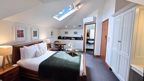 A bedroom with a large bed, a bedside table with a lamp, a small table with a vase, and a view of the kitchen through an open door. - Sandstone Suites (Port Fairy)