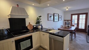 A modern kitchen with a black countertop, white cabinets, and a built-in dishwasher. There is a dining area with a table and chairs, and a dark-colored sofa in the living room. - Sandstone Suites (Port Fairy)