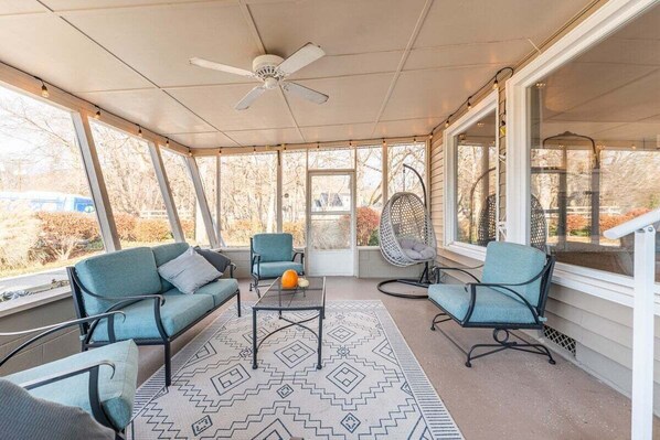 A covered patio with seating, a ceiling fan, and large windows.