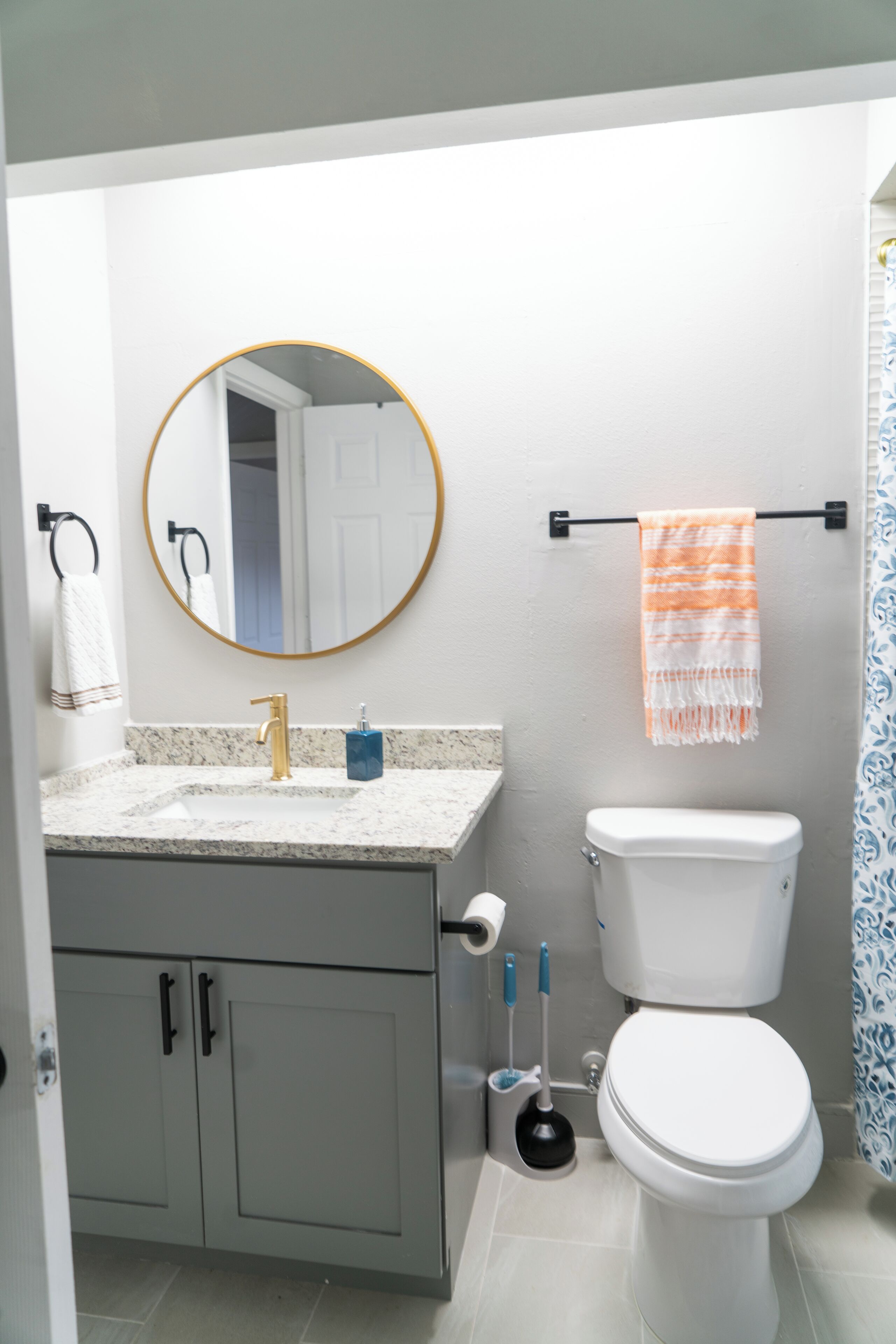 Une salle de bain avec un lavabo blanc, un robinet doré et un miroir rond. On y trouve des toilettes avec une cuvette blanche et un rideau de douche bleu.