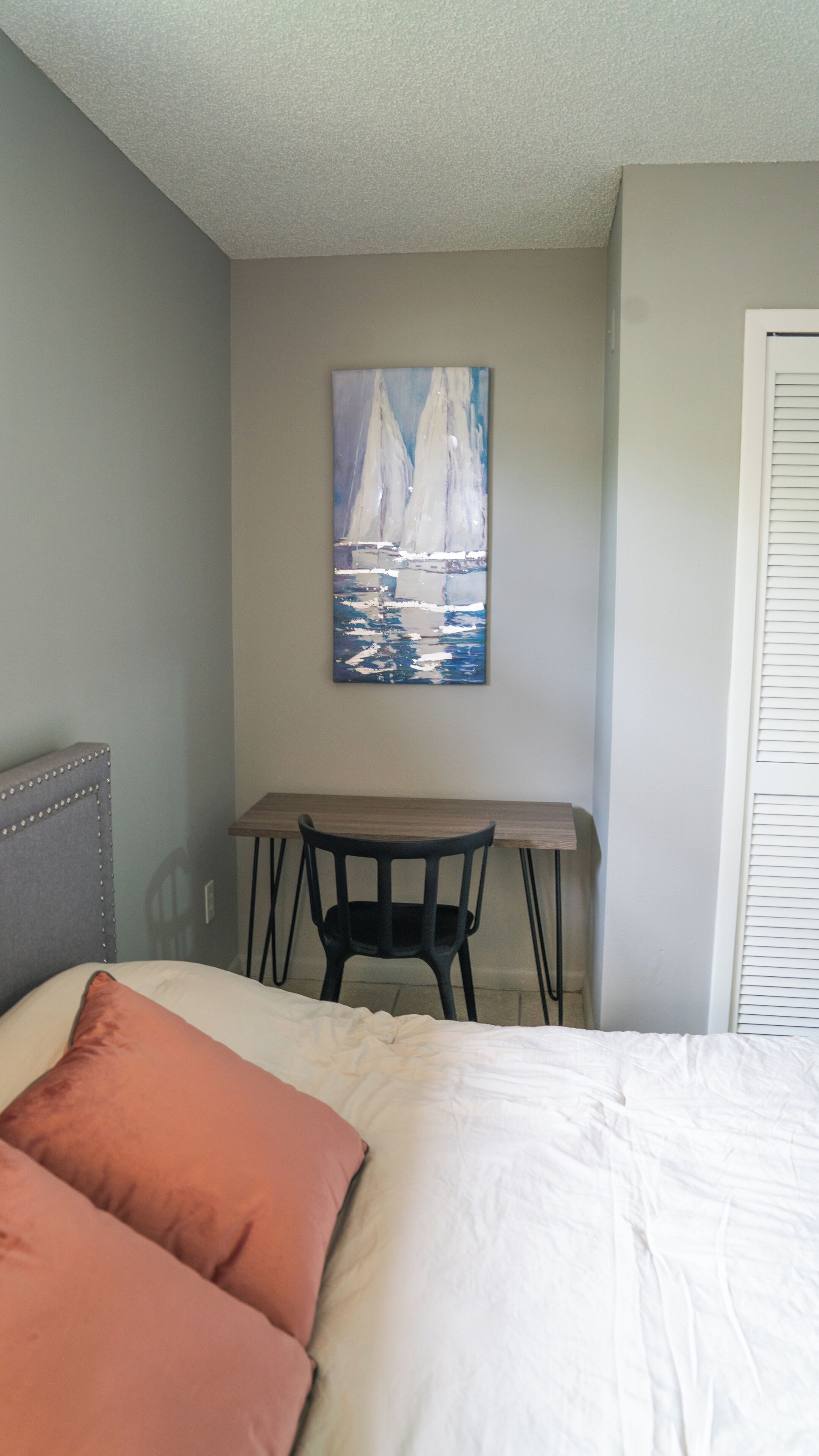 A bedroom with a bed, two pillows, a desk, a chair, and a painting on the wall.