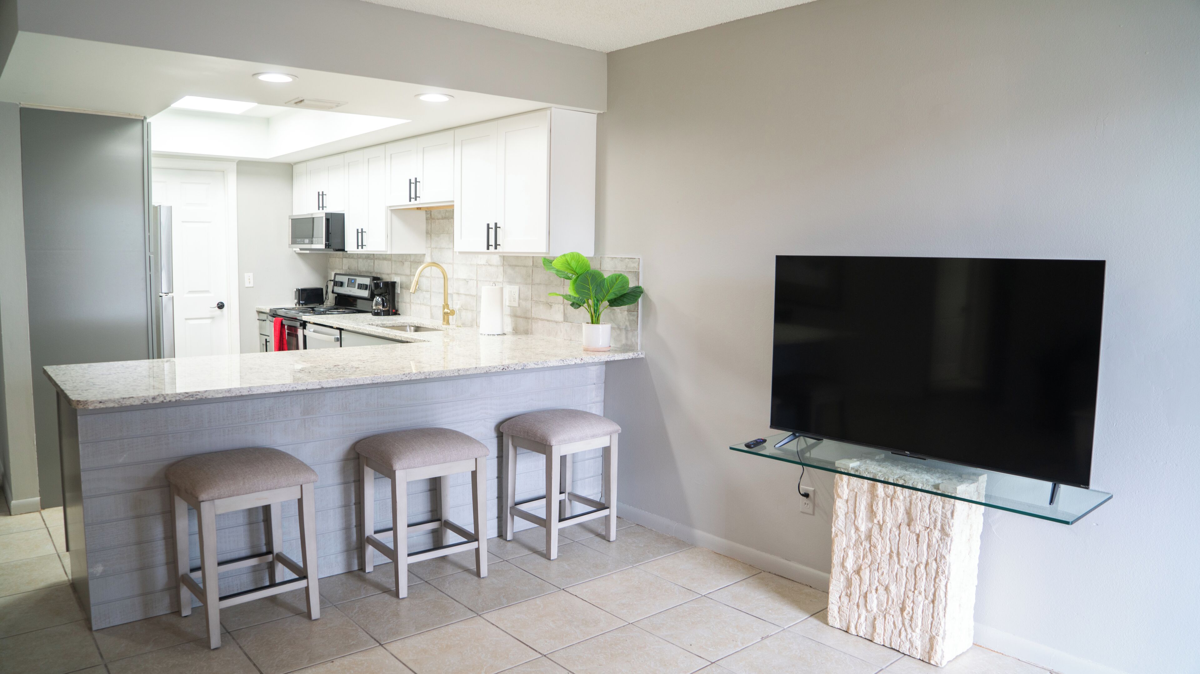 A modern kitchen with a large island, bar stools, a flat-screen TV mounted on a glass shelf, and a potted plant.