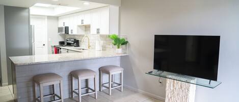 A modern kitchen with a large island, bar stools, a flat-screen TV mounted on a glass shelf, and a potted plant.