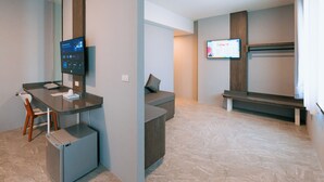 Een moderne hotelkamer met een bureau, een kleine koelkast, een zithoek met een bankje en een flatscreen televisie aan de muur.