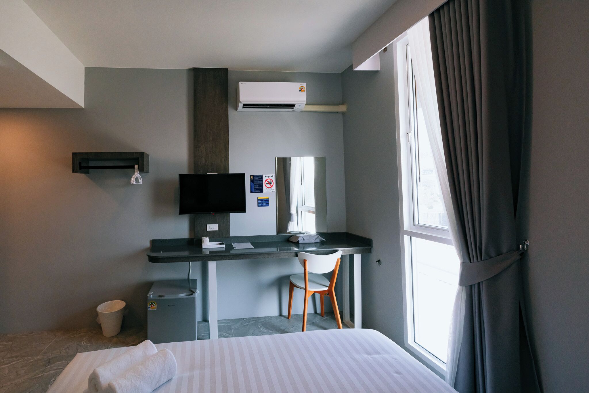 A hotel room with a bed, a desk with a computer, a chair, a small trash bin, and a window with curtains.
