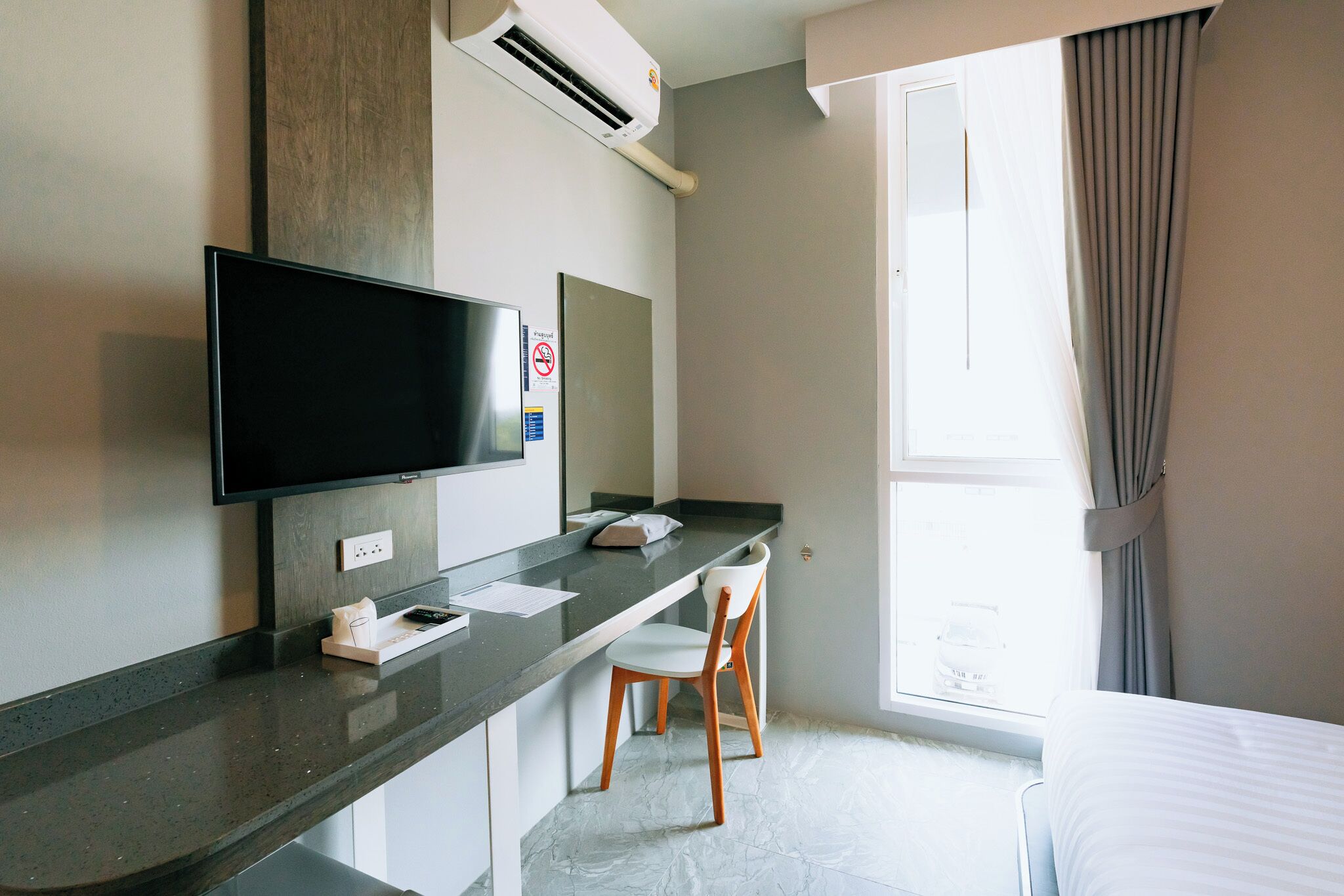 A hotel room with a flat-screen TV mounted on the wall, a small desk with a chair, a bed, and a window with curtains.