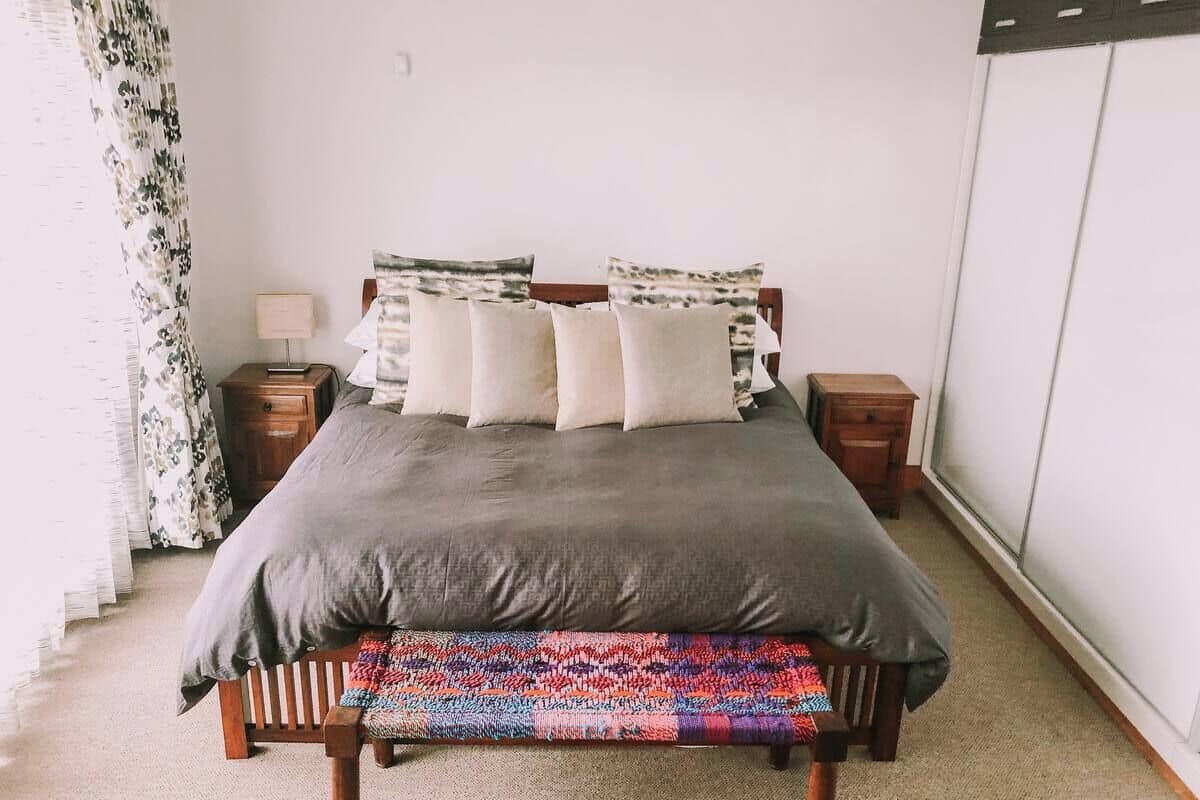Cama bem arrumada com colcha cinza e várias almofadas, um banquinho de madeira com assento colorido trançado, uma mesinha de cabeceira com abajur, e uma janela com cortinas.