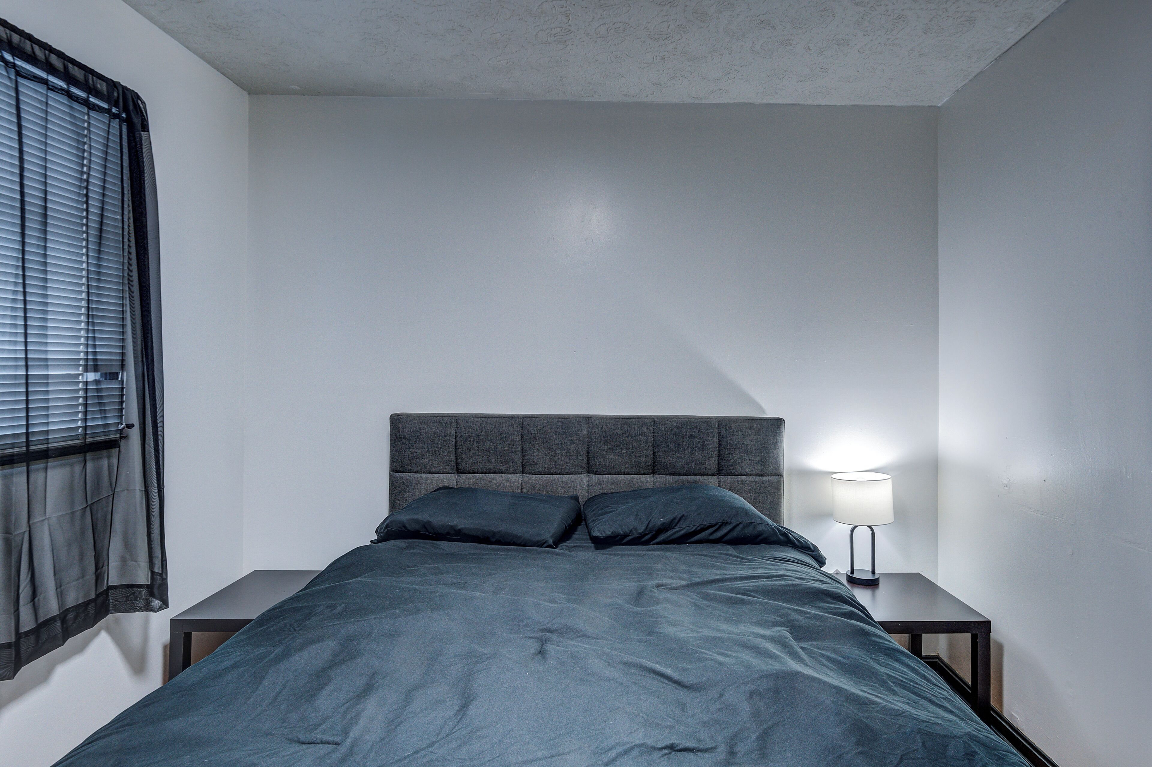 A bedroom with a bed, two pillows, a nightstand with a lamp, and a window with blinds.