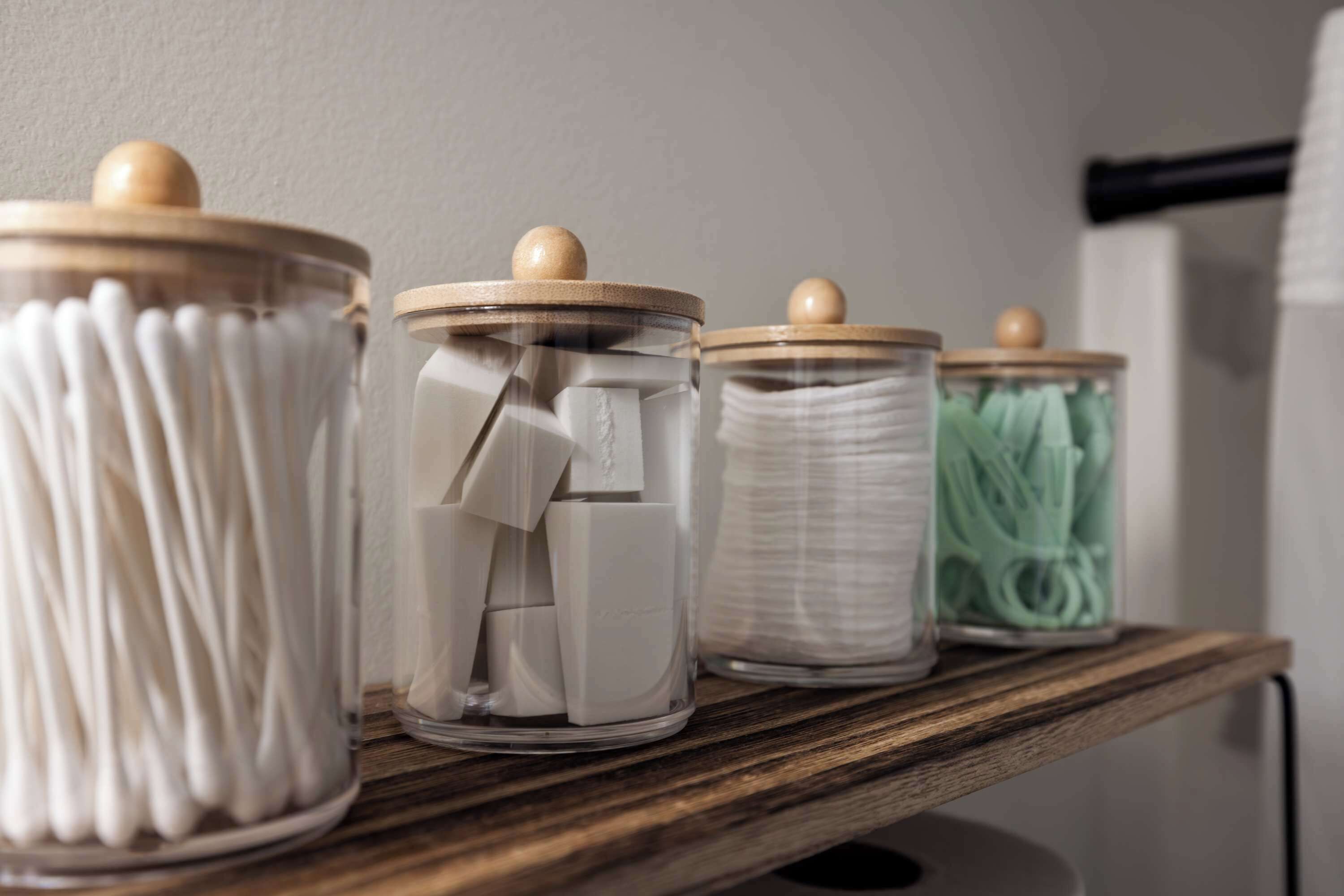 A wooden shelf with three glass jars containing cotton swabs, cotton buds, and a green container.
