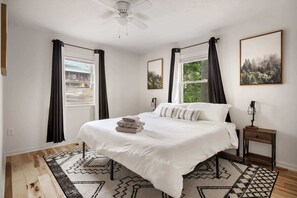 Une chambre à coucher avec un grand lit, deux fenêtres avec des rideaux, un ventilateur de plafond et une table de chevet avec une lampe.