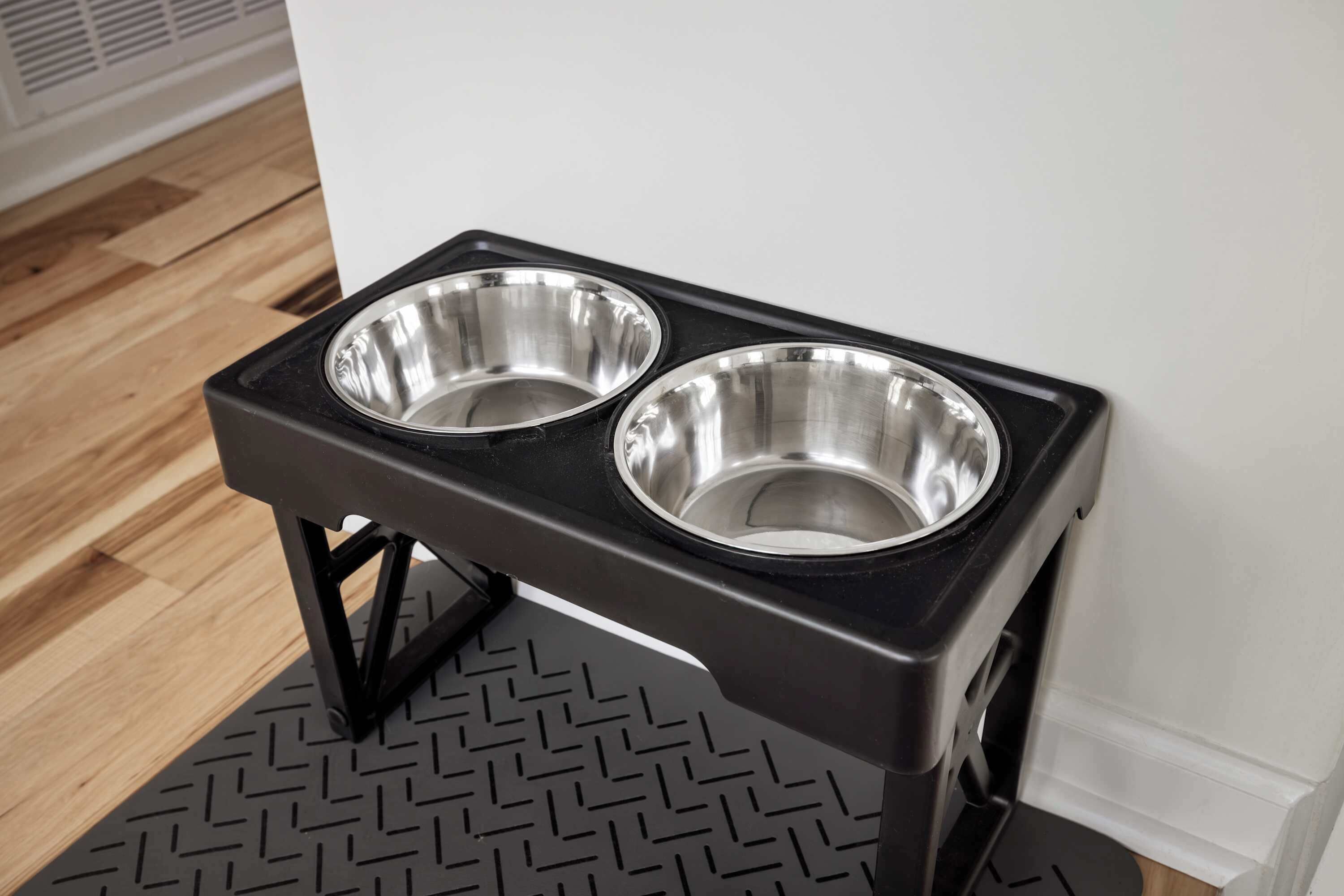 A black pet feeding station with two stainless steel bowls against a white wall.